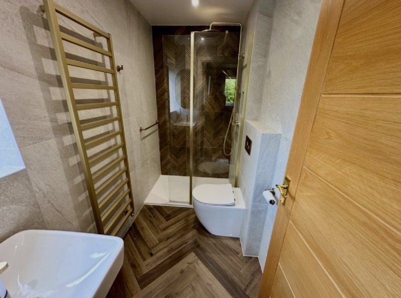Bathroom design and installation by Richard & Richard’s, featuring a walk-in shower, brushed brass fittings, heated towel rail, fitted WC area, large format wall tiles and wood-effect herringbone flooring. Designed, supplied and installed from our Stockpo
