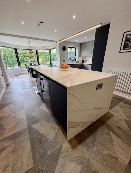 Open-plan kitchen design and installation by Richard & Richard’s, featuring a large statement island, marble-effect waterfall worktop, breakfast bar seating, handleless kitchen furniture, modern pendant lighting and patterned wood-effect flooring. Designe